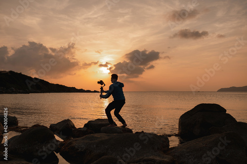 Silhouette of man on rocks in seashore holding a gimbal stabilizer to shot the beautiful landscape at sunset