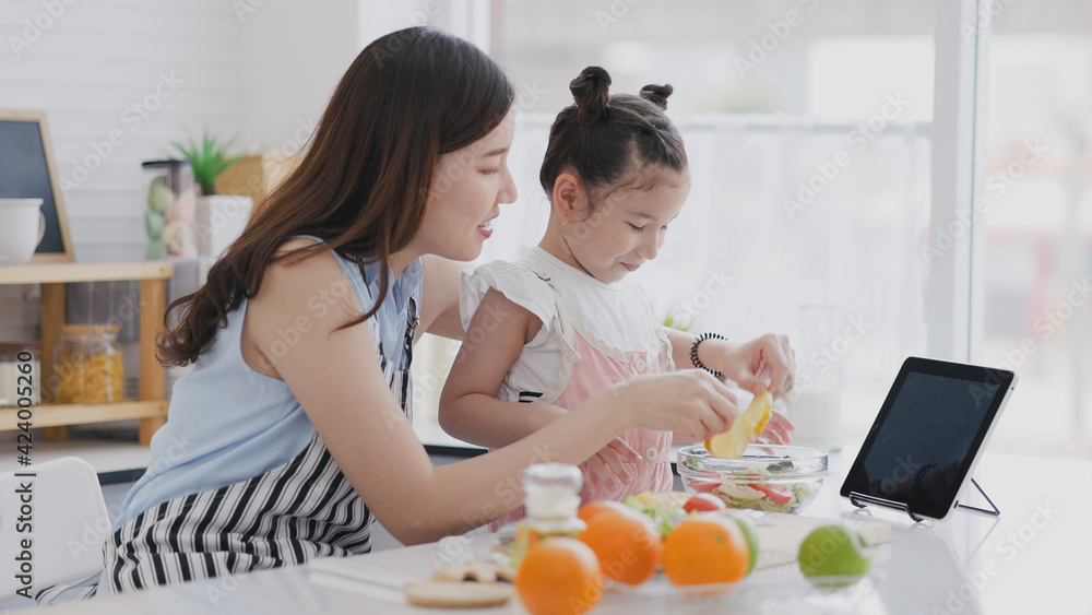 Family single mother and daughter enjoy and happy cooking in clean kitchen. Parents help children make fruit and vegetable salads from Video tutorial from tablet. Learn cook homemade from video call. Family single mother and daughter enjoy and happy cooking in clean kitchen. Parents help children make fruit and vegetable salads from Video tutorial from tablet. Learn cook homemade from video call.