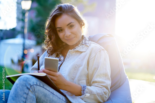 Backlit portrait of attractive female student with modern mobile phone and textbook for studying looking at camera during sunny day at nature, charming blogger with smartphone for networking posing