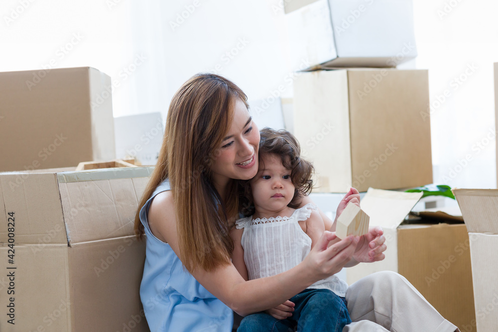 Happy small family while moving into new home. Young Asian mother and her little toddler girl on mother’s lap holding wooden home while moving in new home with cardboard boxes on background Happy small family while moving into new home. Young Asian mother and her little toddler girl on mother’s lap holding wooden home while moving in new home with cardboard boxes on background