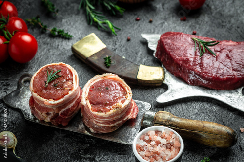 Raw pork medallion steaks wrapped in bacon served on old meat butcher on dark concrete background with spices salt and pepper. Long banner format, top view