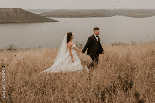 A loving couple wedding newlyweds in a white dress and a suit walk hug kissing on the tall grass in the summer autumn field on the mountain above the river. sunset. bakota ukraine photo