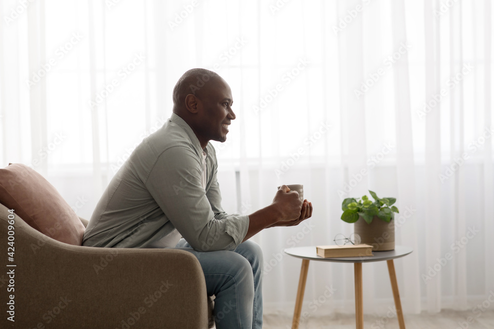 Cozy morning. Happy african american mature man relaxing on armchair and drinking coffee against window