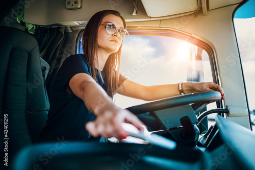 Portrait of beautiful young woman professional truck driver sitting and driving big truck. She is dangerously trying to take smart phone while driving. photo