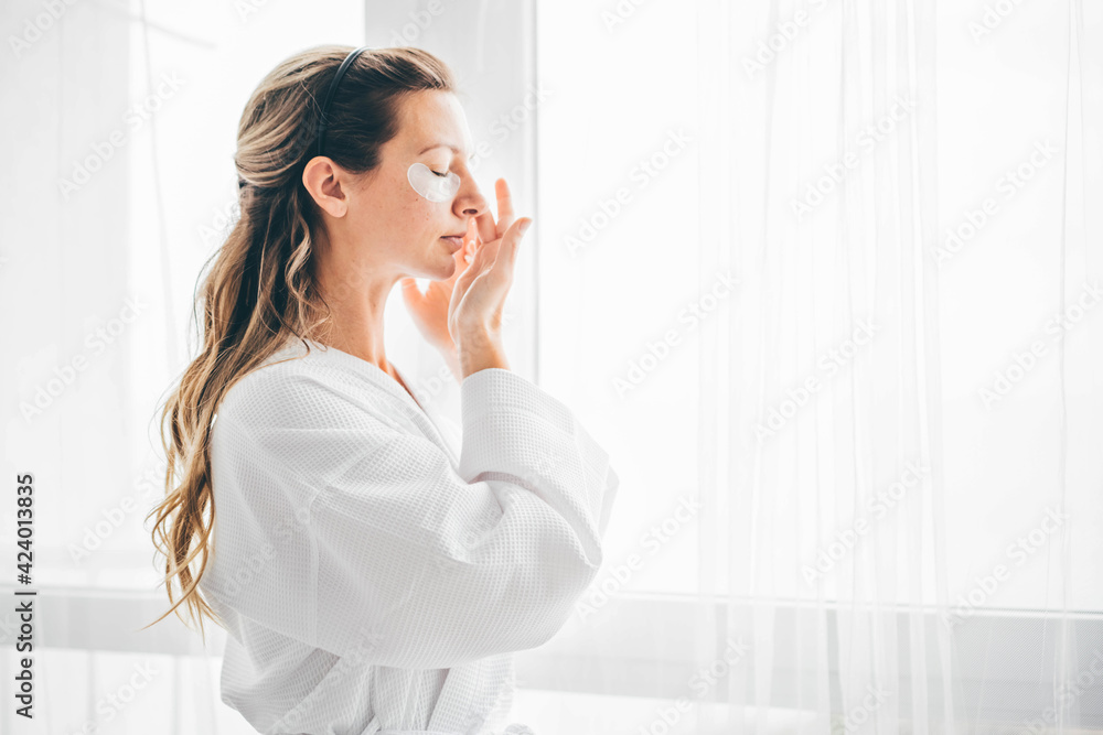 Attractive mature woman in white bathrobe puts anti aging eye patch on face taking care of skin in light modern hotel room close view. Attractive mature woman in white bathrobe puts anti aging eye patch on face taking care of skin in light modern hotel room close view.