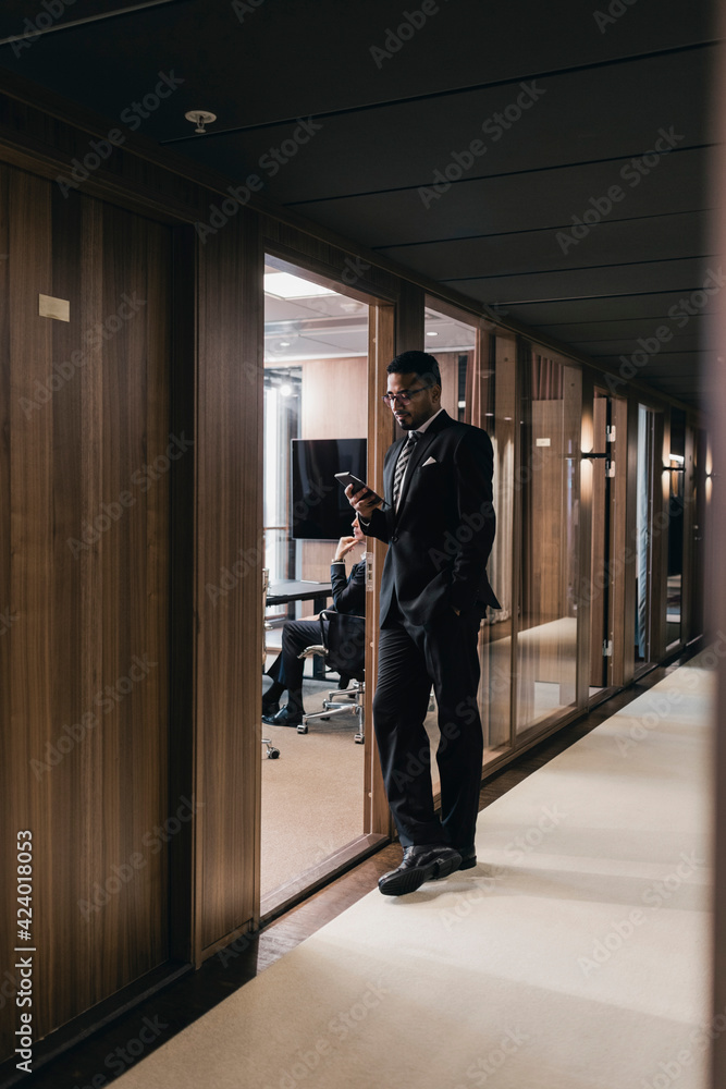 Full length of businessman using smart phone while leaning on board room doorway at office Full length of businessman using smart phone while leaning on board room doorway at office
