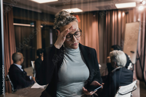 Worried mature businesswoman touching forehead while looking at smart phone against colleagues in board room