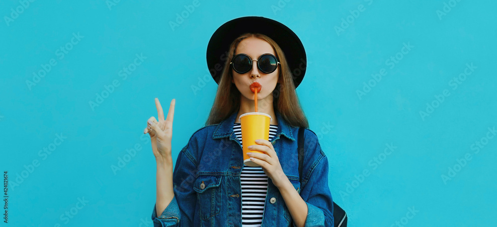 Portrait close up of young woman drinking a juice wearing a black round hat, denim jacket on a blue background