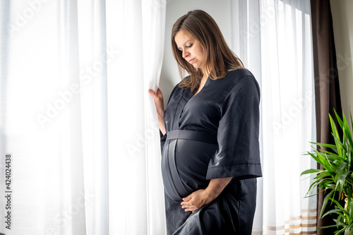 Young pregnant woman looking out of the window and enjoying lovely moment of her first pregnancy