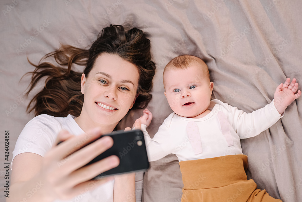 Top view of beautiful mum and baby daughter lying on bed have fun use cell phone watch cartoons, mom with infant girl making online call vlog. Parental control, modern tech, younger generation concept Top view of beautiful mum and baby daughter lying on bed have fun use cell phone watch cartoons, mom with infant girl making online call vlog. Parental control, modern tech, younger generation concept