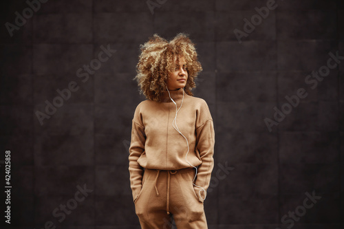 Hip hop girl in tracksuit standing with hands in her pockets, listening music and looking away. photo