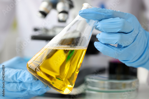 Scientist chemist holding glass flask with yellow liquid in front of microscope closeup