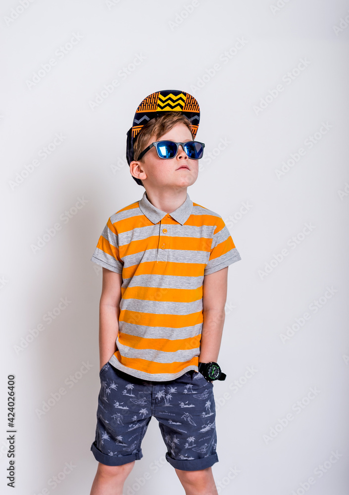 Fototapeta premium Funny blond child in blue glasses, cap, striped shirt. fashionable little boy in sunglasses. stylish kid in yellow shoes. summer fashion children. Summer vacation