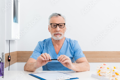 rehabilitologist in eyeglasses holding pen while sitting near clipboard at workplace photo