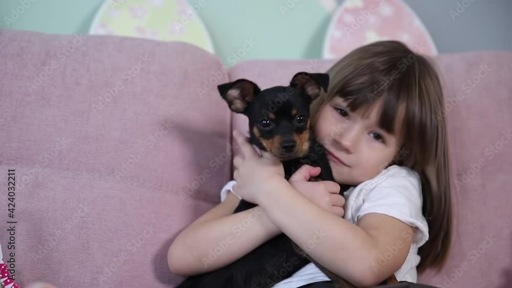 Adorable little girl hug her pet at home on sofa. Kid preparing for Easter at home during quarantine. Cute dog Adorable little girl hug her pet at home on sofa. Kid preparing for Easter at home during quarantine. Cute dog