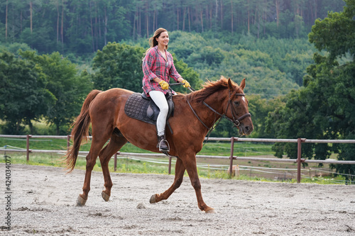 Young woman in shirt riding on brown horse, blurred trees background