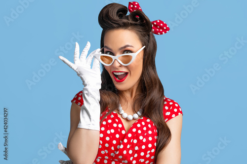 Portrait of excited pin up woman in polka dot dress, headband and gloves touching her sunglasses on blue background