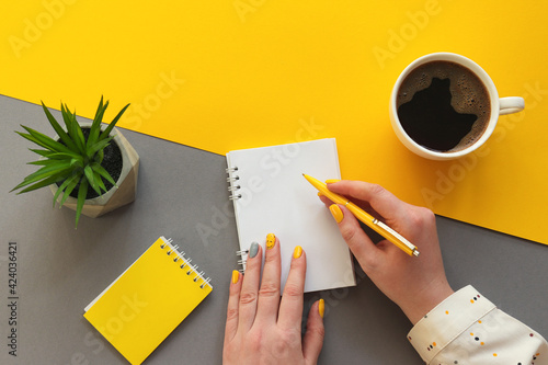 Female hands writing on notepad. Office table desk trendy yellow and grey colors. Workplace with notepads, cup of coffee and succulent plant. Flat lay, top view