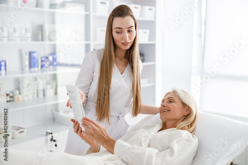 Photo of senior woman in white bathrobe laying on daybed and checking results of skin care treatment while cosmetologist holding looking-glass