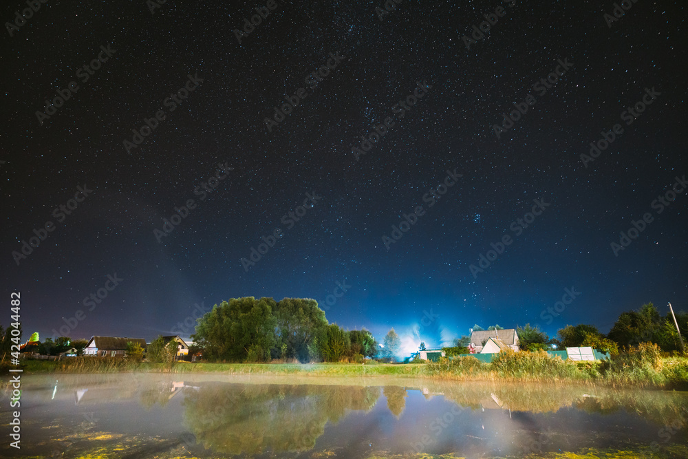 Evaporation Over River Lake Near Houses In Village. Milky Way Galaxy In Night Starry Sky Above Lake River Landscape At Night.