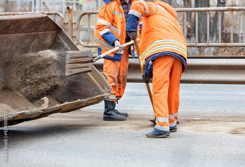 Road workers scrape off accumulated sand between lanes of the road with shovels and load it into a metal grader bucket.