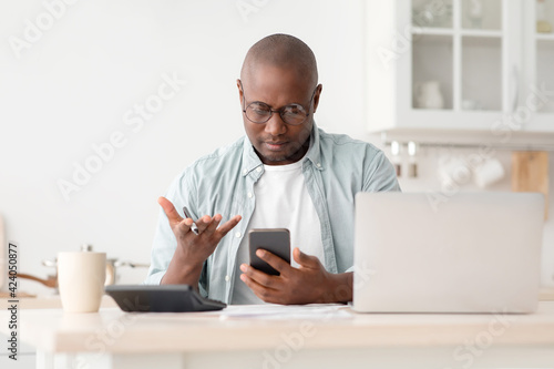 Financial problem. Angry african american man calculating utility charges and bills, using calculator and phone