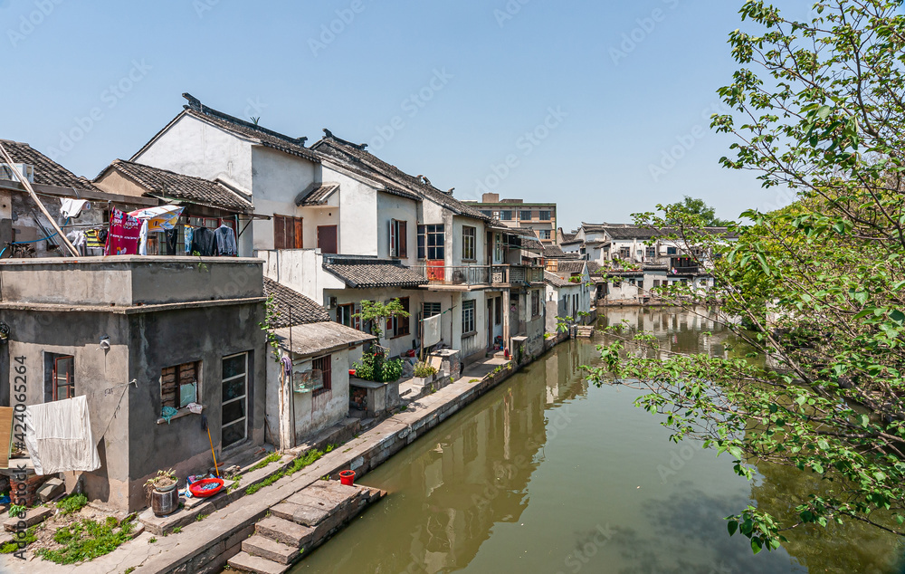 Tongli, China - May 2, 2010: Greenish water in canal with generic architecture white houses on one side under light blue sky. Some green foliage and extra colors by drying laundry added.