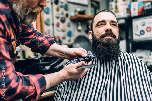 The barber trims the beard of the customer in his barber shop. Trendy and stylish beard styling and cut.
