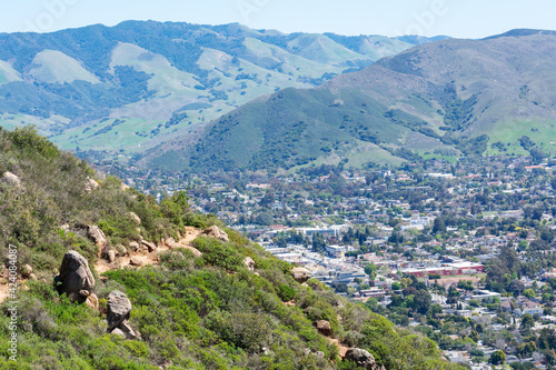 Aerial view of Bishop Peak trail surrounded by chaparral on the hill. Background San Luis Obispo area sprawl and scenic green mountains of Santa Lucia Range.