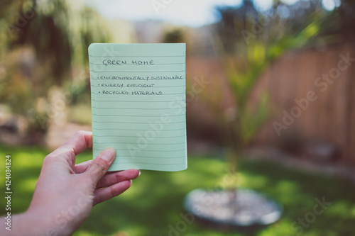 eco-friendly construction, hand holding house icon and green home elements list under the sun with garden bokeh bakcground. photo