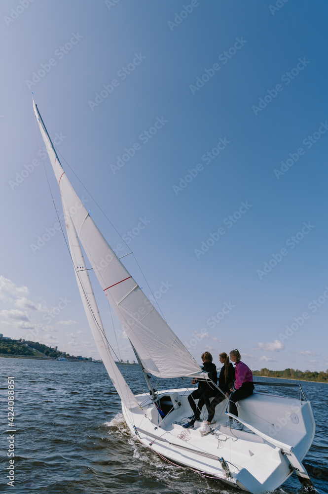 Obraz premium A beautiful white racing single-masted yacht is sailing against a beautiful river landscape with a blue sky. A man and two girls are on board.