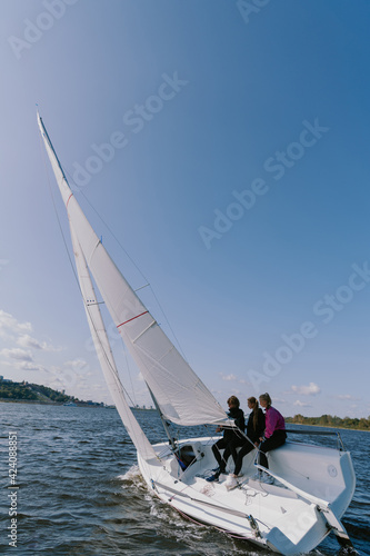 A beautiful white racing single-masted yacht is sailing against a beautiful river landscape with a blue sky. A man and two girls are on board.