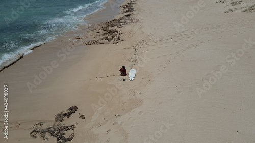 Young Female surfer sitting on the beach looking thinking to go surf at the waves of the rolling Pacific Ocean at a sandy beach near Exmouth, Western Australia.