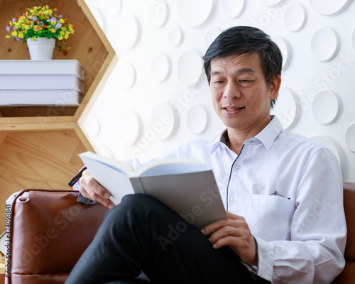 Asian old middle aged confident black and gray hair male businessman wears white long sleeve shirt black pants glasses in pocket sit on brown leather couch having problem to read white book in hands photo