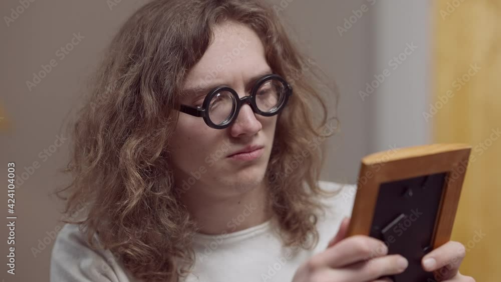Sad young Caucasian man in retro eyeglasses with long curly hair looking at photograph and crying. Close-up portrait of desperate frustrated depressed guy at home indoors. Nostalgia concept. Sad young Caucasian man in retro eyeglasses with long curly hair looking at photograph and crying. Close-up portrait of desperate frustrated depressed guy at home indoors. Nostalgia concept.