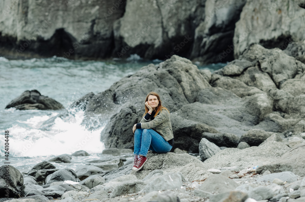 young woman in jeans and red sneakers sits on rocks near the stormy oceans with waves. local tourism, solo travel. mental health communication with nature.