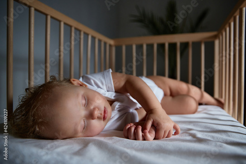 Cute baby sleeps at night in a cradle for babies without a canopy and bumpers. Safe sleep in a dark, spacious room with a cot and a flower. Interior of the bedroom and children's room