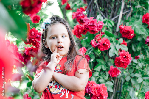 portrait of a plump, cheeky five-year-old girl with her arms crossed and her finger at her mouth against the background of a park and rose bushes.