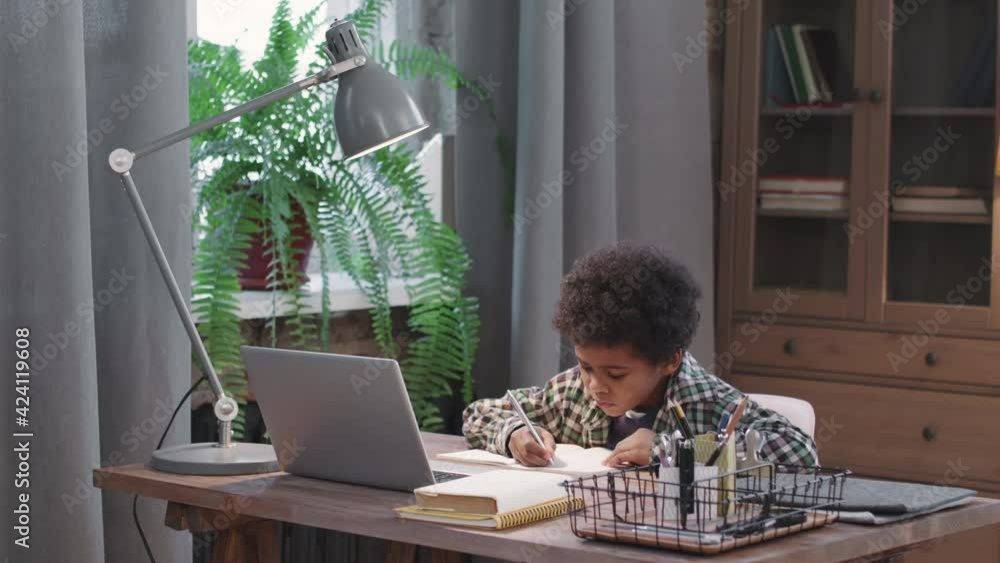 Panning medium shot with slowmo of smart and cute 8-year-old afro kid sitting at desk in living room studying online via laptop, writing in copybook Panning medium shot with slowmo of smart and cute 8-year-old afro kid sitting at desk in living room studying online via laptop, writing in copybook