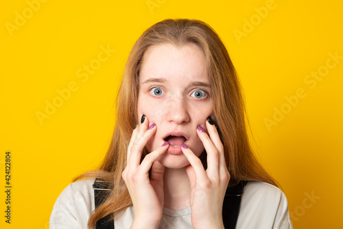 Nervous scared girl grabs face and looks with worried expression at camera, sees phobia, afraid of speaking, on yellow background. Human reaction concept.