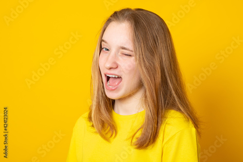 Childish and playful teenage girl winking at camera, keeping mouth wide opened on yellow background with copy space for your text or promotional information. © Eugene