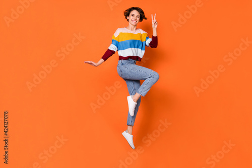 Photo of charming pretty young woman wear striped sweater showing v-sign jumping high isolated orange color background