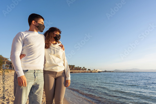 Mixed race heterosexual couple stand hugging on sea shore at beach wearing protective mask for restriction from Covid-19 pandemic looking towards the horizon at sunset. Young people trusting future