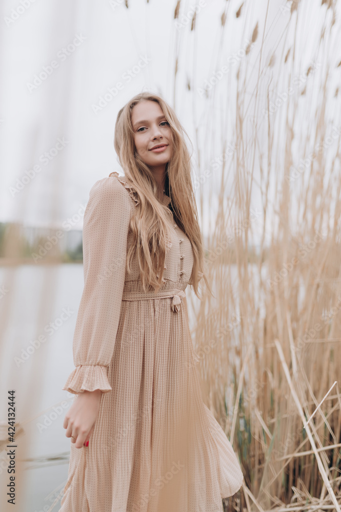 Naklejka premium a beautiful girl with brown hair in a beige dress stands in the tall reeds by the water