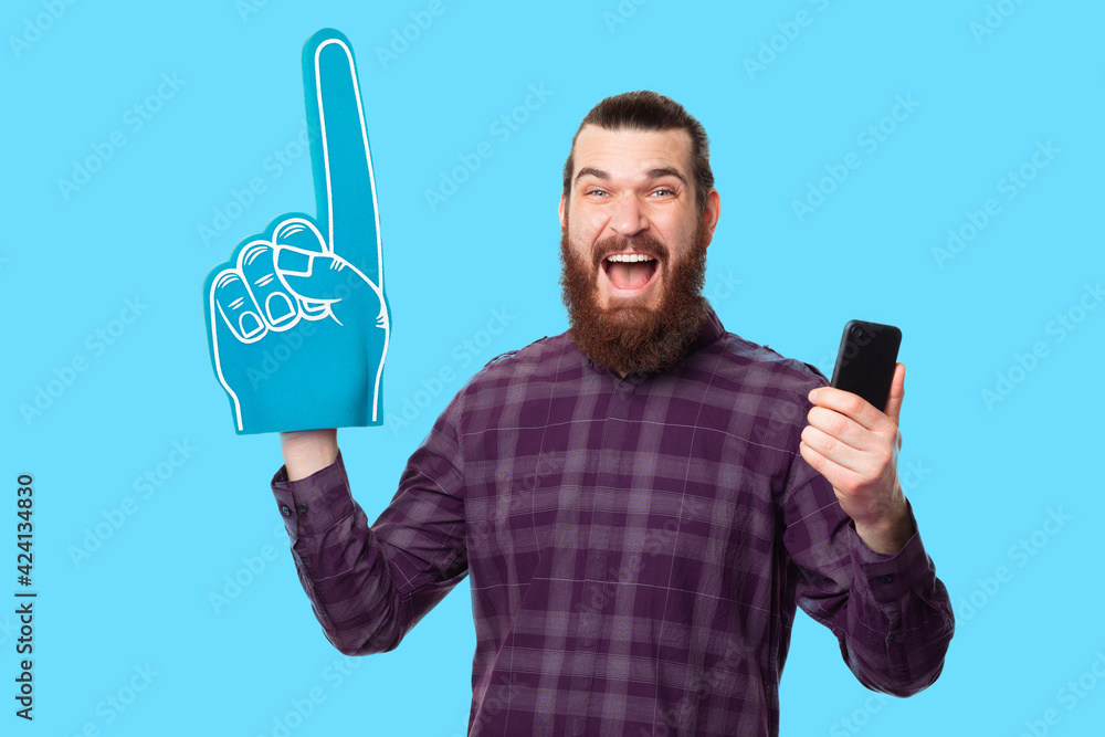 Photo of screaming amazed bearded man pointing with fan glove and holding smartphone, supporting team Photo of screaming amazed bearded man pointing with fan glove and holding smartphone, supporting team