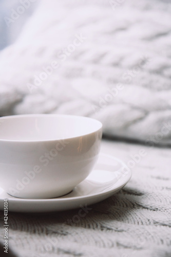 Large white ceramic cup on a saucer on a knitted nude blanket. Cozy home background. Still life from home interior