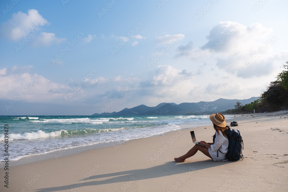 A female traveler using and working on laptop computer while sitting on the beach A female traveler using and working on laptop computer while sitting on the beach