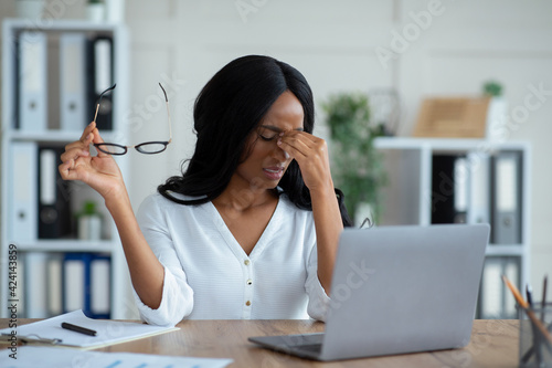 Black young businesswoman having tired irritated eyes, sitting at desk with laptop, exhausted from online work in office