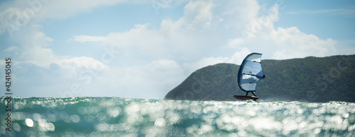 A man wing foils at sea using a hand held inflatable wing, riding a hydrofoil surfboard.  Turquoise sea, cloudy blue sky. 