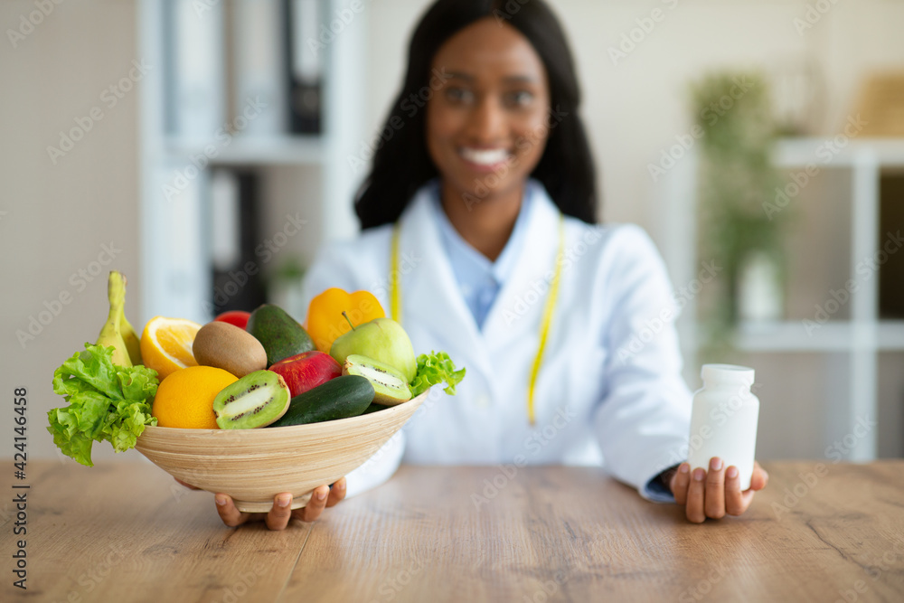 Happy black dietitian offering choice between fresh products and tablets at clinic Happy black dietitian offering choice between fresh products and tablets at clinic